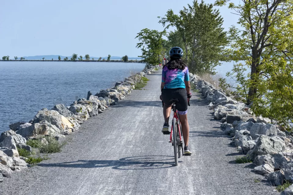 View from behind of woman wearing helmet on bicycle riding on concrete causeway with water on either side.